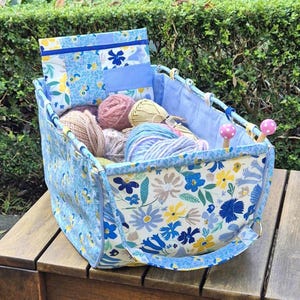 May include: A rectangular fabric yarn storage basket featuring a blue, yellow, and white floral pattern. The basket contains several balls of yarn in various colours and includes a matching zippered pouch and two wooden knitting needles with pink toppers.