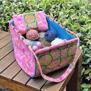 May include: A pink and teal fabric yarn tote bag with a matching pouch. The tote has a blue interior and is filled with various colors of yarn. The exterior features a tree and floral pattern. A pink strap is attached to the tote.