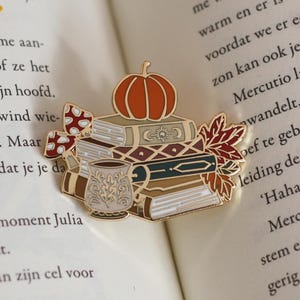 Op de afbeelding: Een goudkleurige emaille pin met een stapel boeken, een pompoen, herfstbladeren en een decoratieve mok. De pin heeft een herfstthema, ideaal voor boekenliefhebbers.