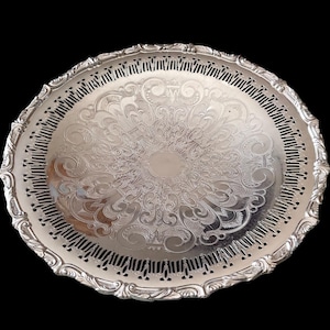 May include: A silver-coloured, round serving tray with an ornate, floral-inspired pattern. The tray has a decorative, scalloped edge with small cut-out details. The background is solid black.