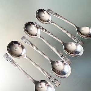 May include: A collection of six silver spoons. Each spoon has a rounded bowl and a long handle with a decorative floral design at the end. The spoons are arranged on a reflective surface, with their polished surfaces reflecting the surrounding environment.