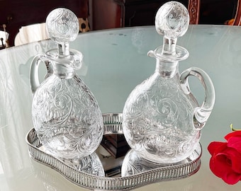 May include: Two clear glass decanters with etched floral designs, sitting on a silver tray with a mirrored surface. A single white rose is on the table next to the tray.