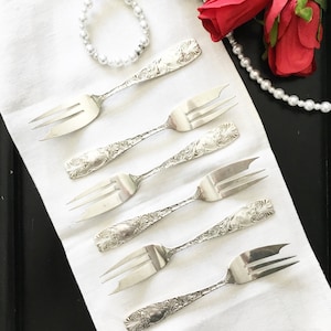 May include: A set of six silver cake forks with ornate handles, laid out on a white linen cloth. The forks are arranged in a staggered pattern, with the handles pointing towards the top of the image.