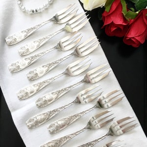 May include: A set of ten silver forks with ornate designs. The forks are laid out on a white cloth surface.