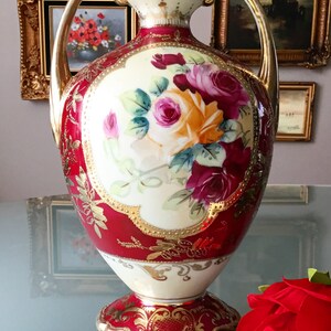 Op de afbeelding: Een rode en gouden porseleinen vaas met een bloemenmotief. De vaas heeft twee handvatten en een gegolfde basis. De bloemen zijn geschilderd in tinten roze, rood en geel.