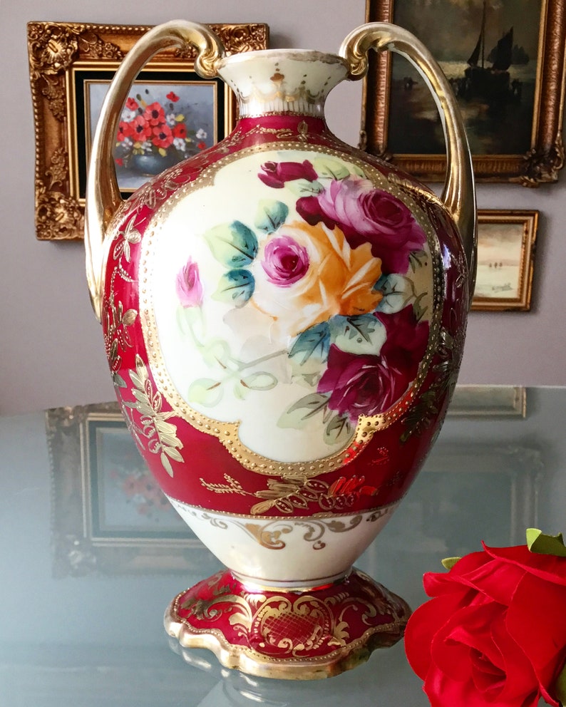 Op de afbeelding: Een rode en gouden porseleinen vaas met een bloemenmotief. De vaas heeft twee handvatten en een gegolfde basis. De bloemen zijn geschilderd in tinten roze, rood en geel.