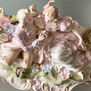 Op de afbeelding: Een porseleinen beeldje van een stel in een romantische omhelzing, omringd door delicate bloemen. De man draagt een roze en gouden pak, terwijl de vrouw een roze en witte jurk draagt. Het beeldje staat op een groene en witte basis.