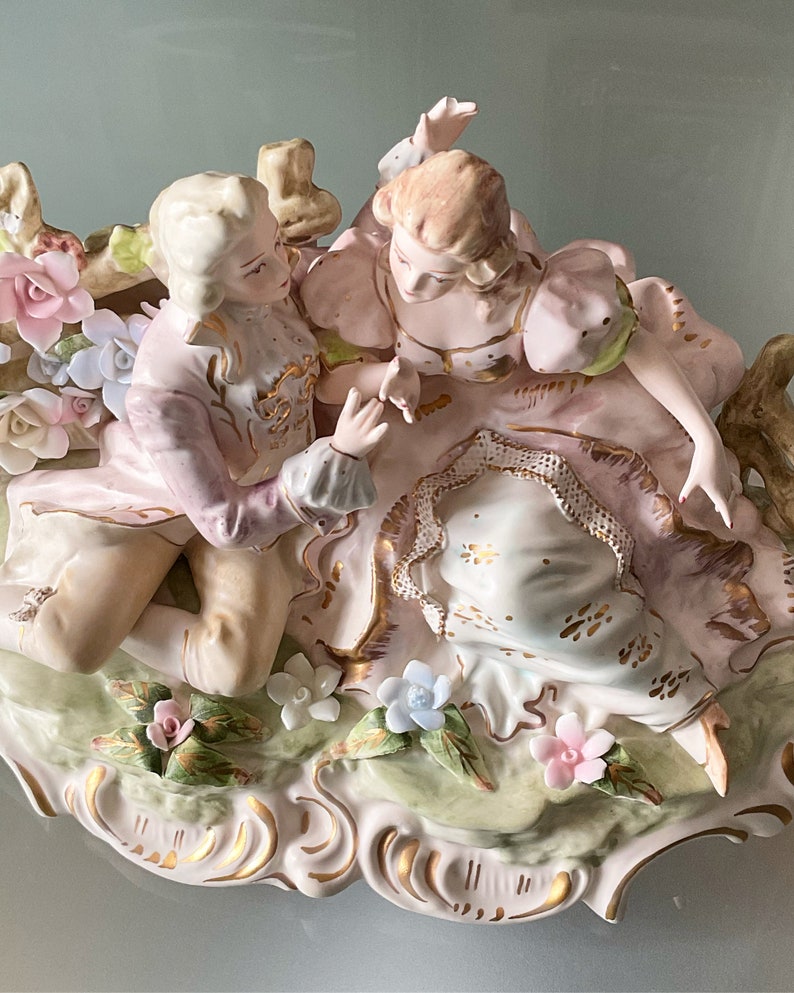 Op de afbeelding: Een porseleinen beeldje van een stel in een romantische omhelzing, omringd door delicate bloemen. De man draagt een roze en gouden pak, terwijl de vrouw een roze en witte jurk draagt. Het beeldje staat op een groene en witte basis.