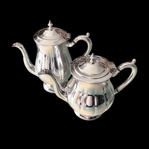 May include: Two silver teapots with ornate handles and lids. The teapots are polished and reflect light.