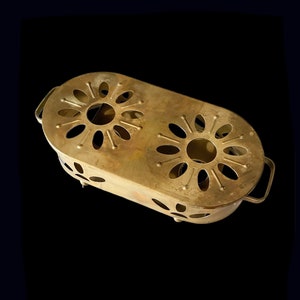 May include: A brass tea cosy with two cut-out flower designs. The cosy has two handles and sits on four small feet.