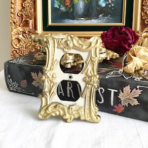 May include: A gold-coloured decorative outlet cover with an ornate, rococo-style frame. The cover is white and has a single outlet.