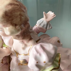 Op de afbeelding: Een porseleinen beeldje van een vrouw in een roze jurk met gouden accenten. Ze zit met haar rechterarm omhoog en haar hand uitgestoken. Haar haar is bruin en haar jurk heeft een groene zoom.