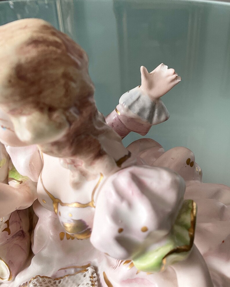 Op de afbeelding: Een porseleinen beeldje van een vrouw in een roze jurk met gouden accenten. Ze zit met haar rechterarm omhoog en haar hand uitgestoken. Haar haar is bruin en haar jurk heeft een groene zoom.