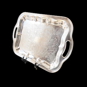 May include: A silver serving tray with detailed floral and scrollwork patterns. The tray has a rectangular shape with a scalloped edge and integrated handles. The tray is displayed against a black background.
