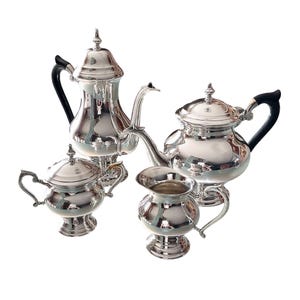 May include: A silver tea set comprising a teapot, creamer, sugar bowl, all with matching designs. The set features black handles and a polished finish.