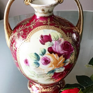 Op de afbeelding: Een rode en gouden porseleinen vaas met twee handvatten en een bloemenontwerp. De vaas is versierd met gouden accenten en heeft een boeket roze en gele rozen.