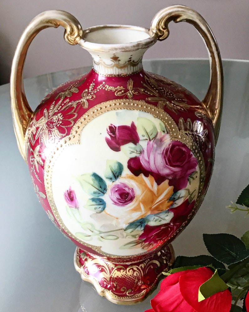 Op de afbeelding: Een rode en gouden porseleinen vaas met twee handvatten en een bloemenontwerp. De vaas is versierd met gouden accenten en heeft een boeket roze en gele rozen.