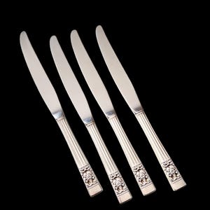 May include: Four silver knives with decorative floral designs on the handles. The blades are smooth and shiny, and the handles have vertical ridges. The knives are arranged diagonally against a black background.