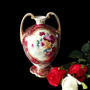 Op de afbeelding: Een rode en gouden porseleinen vaas met een bloemenmotief en twee handvatten. De vaas is versierd met een bloemenpatroon in tinten roze, geel en groen. De vaas staat op een zwart oppervlak met drie rode rozen en &eacute;&eacute;n witte roos.