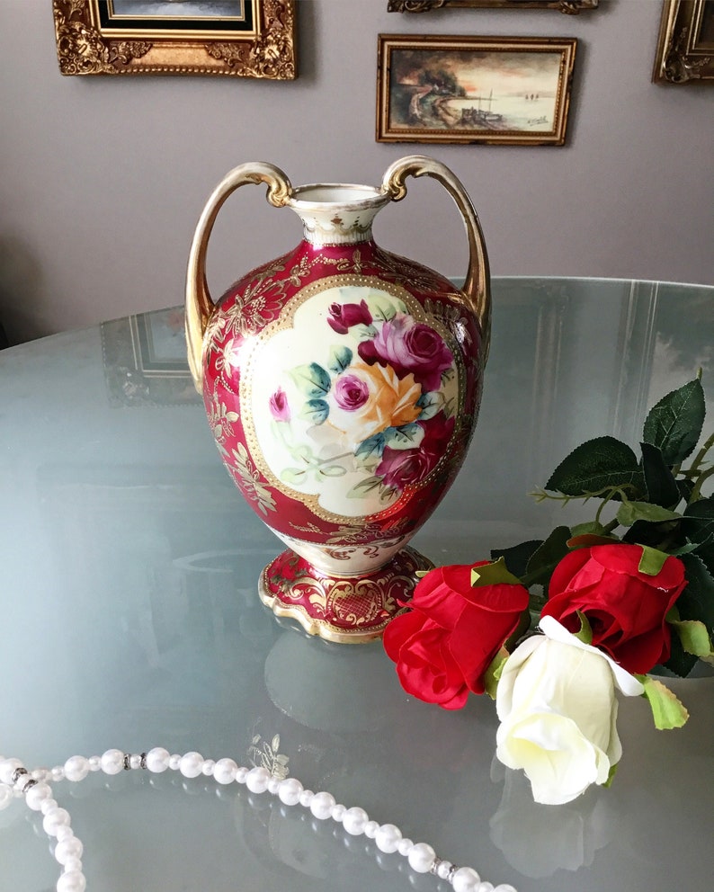 Op de afbeelding: Een rode en gouden porseleinen vaas met bloemenmotieven en twee handvatten. De vaas is versierd met een bloemenpatroon in tinten roze, geel en groen. De vaas staat op een glazen tafel met een parelketting.