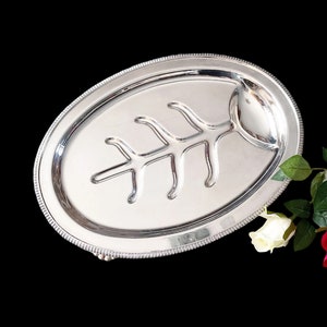 May include: A silver oval serving platter with a decorative leaf design and a small round indentation. The platter has a beaded edge and three feet.