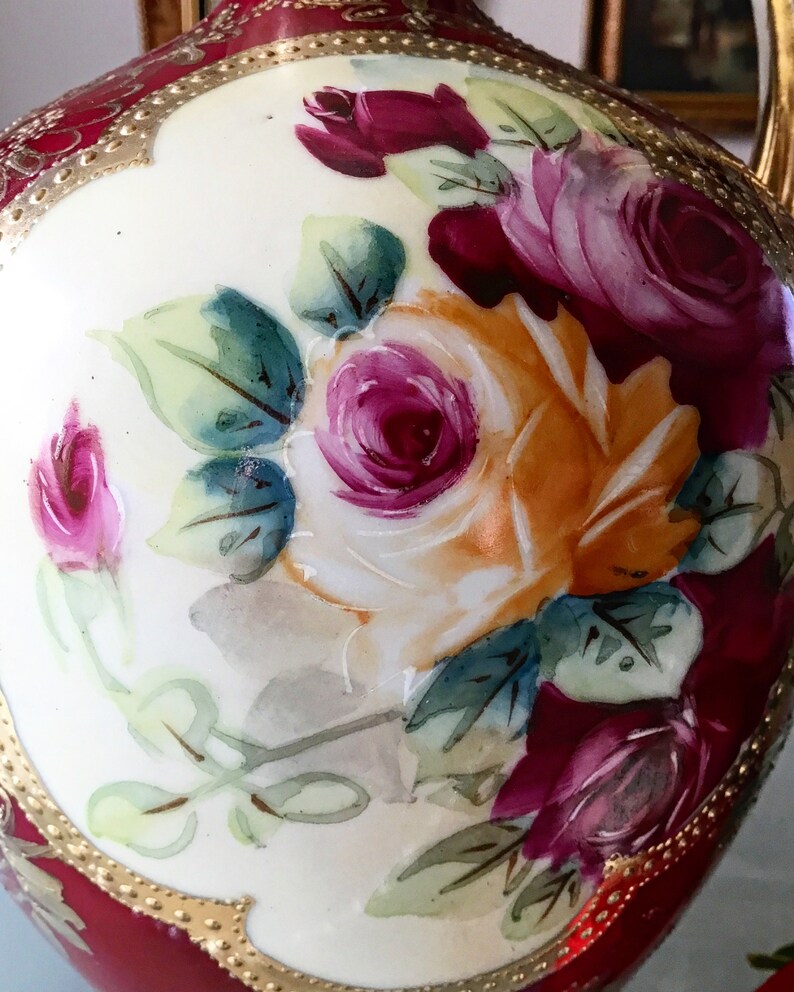 Op de afbeelding: Een vintage porseleinen vaas met een rode en gouden basis en een wit lichaam. De vaas is versierd met handgeschilderde rozen in tinten roze, rood en geel.