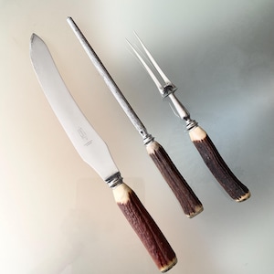 May include: A three-piece carving set consisting of a curved steel knife, a sharpening steel, and a two-pronged carving fork. Each utensil features a textured, dark brown handle with a natural finish and a silver-toned metal bolster.