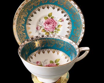 May include: A white teacup and saucer with a teal blue and gold floral design. The cup and saucer have a gold rim and a floral pattern with pink roses.