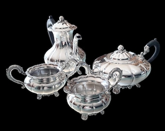 May include: A silver tea set with a teapot, creamer, sugar bowl, and a serving pot. The set has ornate handles and decorative detailing. The teapot has a black handle. The set is displayed against a black background.