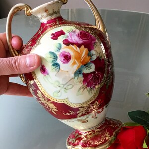 Op de afbeelding: Een rode en gouden porseleinen vaas met een bloemenmotief. De vaas heeft twee handvatten en een gegolfde basis. De bloemen zijn geschilderd in tinten roze, geel en rood.