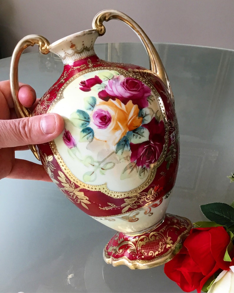 Op de afbeelding: Een rode en gouden porseleinen vaas met een bloemenmotief. De vaas heeft twee handvatten en een gegolfde basis. De bloemen zijn geschilderd in tinten roze, geel en rood.