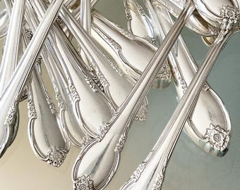 May include: A close-up of a pile of silver flatware with ornate handles. The silverware is arranged in a fan shape, with the handles pointing towards the top of the image. The silverware is shiny and polished, reflecting the light.