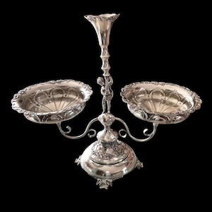 May include: A silver three-tiered serving dish with two bowls and a central vase. The bowls and vase have ornate designs and the base has a decorative floral pattern.