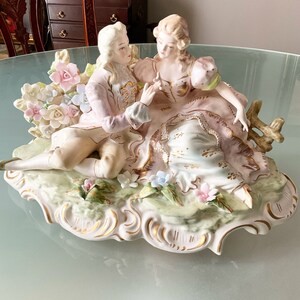 Op de afbeelding: Een porseleinen beeldje van een stel in een romantische omhelzing, versierd met delicate bloemdetails en gouden accenten. Het stel zit op een decoratieve basis met een bloemmotief.