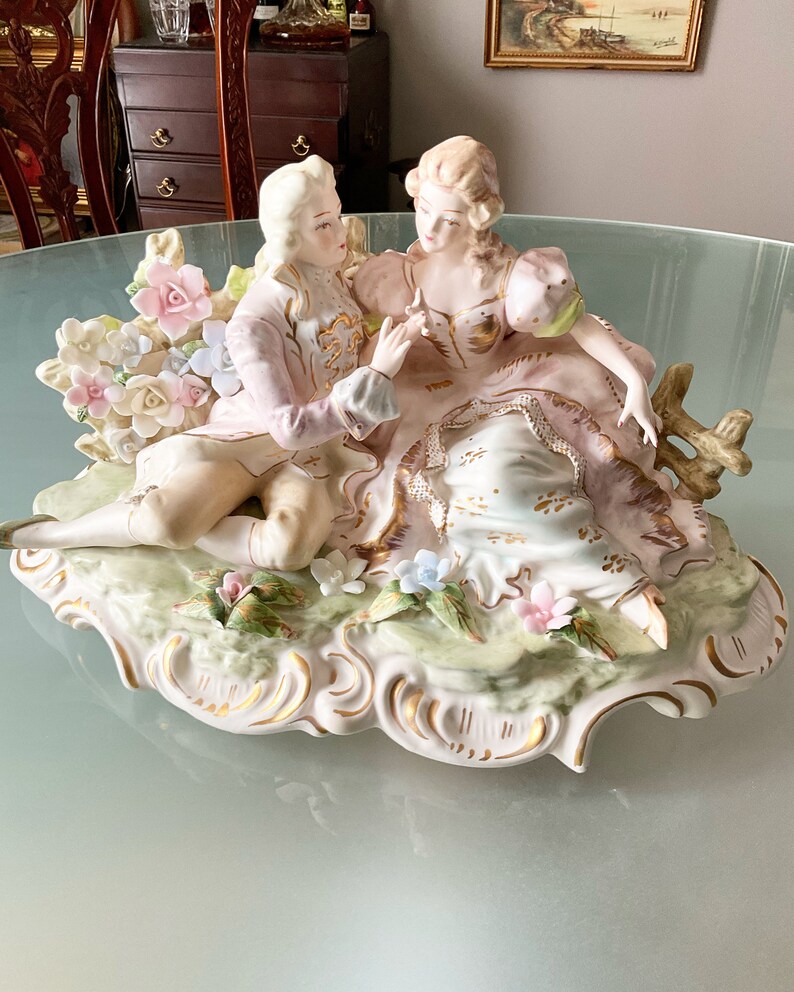 Op de afbeelding: Een porseleinen beeldje van een stel in een romantische omhelzing, versierd met delicate bloemdetails en gouden accenten. Het stel zit op een decoratieve basis met een bloemmotief.
