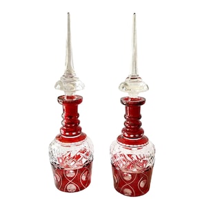 May include: A pair of ruby red glass decanters with clear glass stoppers and intricate cut glass designs. The decanters have a round body and a tall, slender neck.