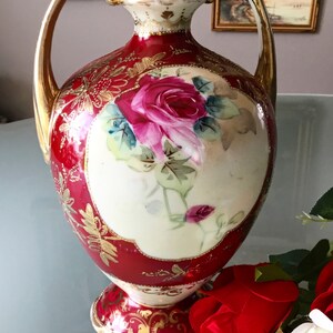 Op de afbeelding: Een rode en gouden porseleinen vaas met twee handvatten en een bloemenontwerp. De vaas heeft een grote roze roos met groene bladeren en gouden accenten.