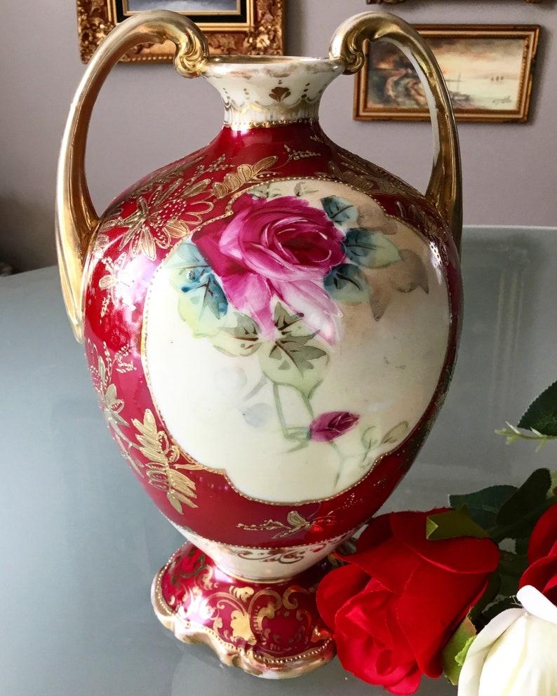 Op de afbeelding: Een rode en gouden porseleinen vaas met twee handvatten en een bloemenontwerp. De vaas heeft een grote roze roos met groene bladeren en gouden accenten.