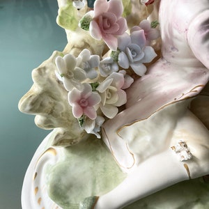 Op de afbeelding: Een porseleinen beeldje van een vrouw in een witte jurk met een roze en groen bloemenmotief. De vrouw zit op een groene en witte basis met gouden accenten. Het beeldje is versierd met delicate bloemen in tinten roze, wit en blauw.