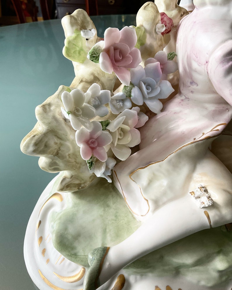 Op de afbeelding: Een porseleinen beeldje van een vrouw in een witte jurk met een roze en groen bloemenmotief. De vrouw zit op een groene en witte basis met gouden accenten. Het beeldje is versierd met delicate bloemen in tinten roze, wit en blauw.