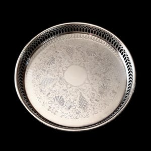May include: A silver-coloured, round serving tray with an ornate, engraved floral design. The tray has a decorative, openwork edge and a plain, circular centre. The background is solid black.