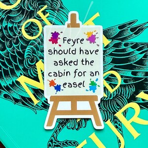 May include: A teal book titled "A Court of Thorns and Roses" by Sarah J. Maas, with a raven illustration. A small easel-shaped sticker with the text "Feyre should have asked the cabin for an easel" is placed on the book.