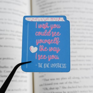 May include: A blue book-shaped sticker with the text "I wish you could see yourself the way I see you" in pink script. A white heart is next to the text. The sticker also includes the text "- THE LOVE HYPOTHESIS".