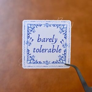 May include: A white square sticker with a blue and white floral border. The text "barely tolerable" is written in blue cursive lettering.