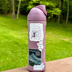 K&ouml;nnte beinhalten: Eine mauvefarbene Owala-Trinkflasche mit Klappdeckel. Die Flasche ist mit Aufklebern mit Text und Illustrationen verziert, darunter ein Hirsch, eine Berglandschaft und S&auml;tze wie "Greetings from the Night Court."