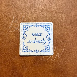 May include: A square sticker with a white background and blue floral border. The words "most ardently" are written in blue script. The sticker is on a brown leather-like surface.