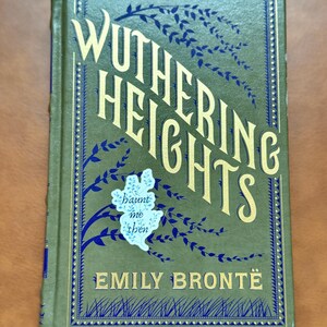 K&ouml;nnte beinhalten: Ein Hardcover-Buch mit dem Titel "Wuthering Heights" von Emily Bront&euml;. Das Cover ist olivgr&uuml;n mit goldfarbenen Buchstaben und blauen floralen Akzenten. Ein kleiner Aufkleber mit den Worten "haunt me then" befindet sich auf dem Cover.