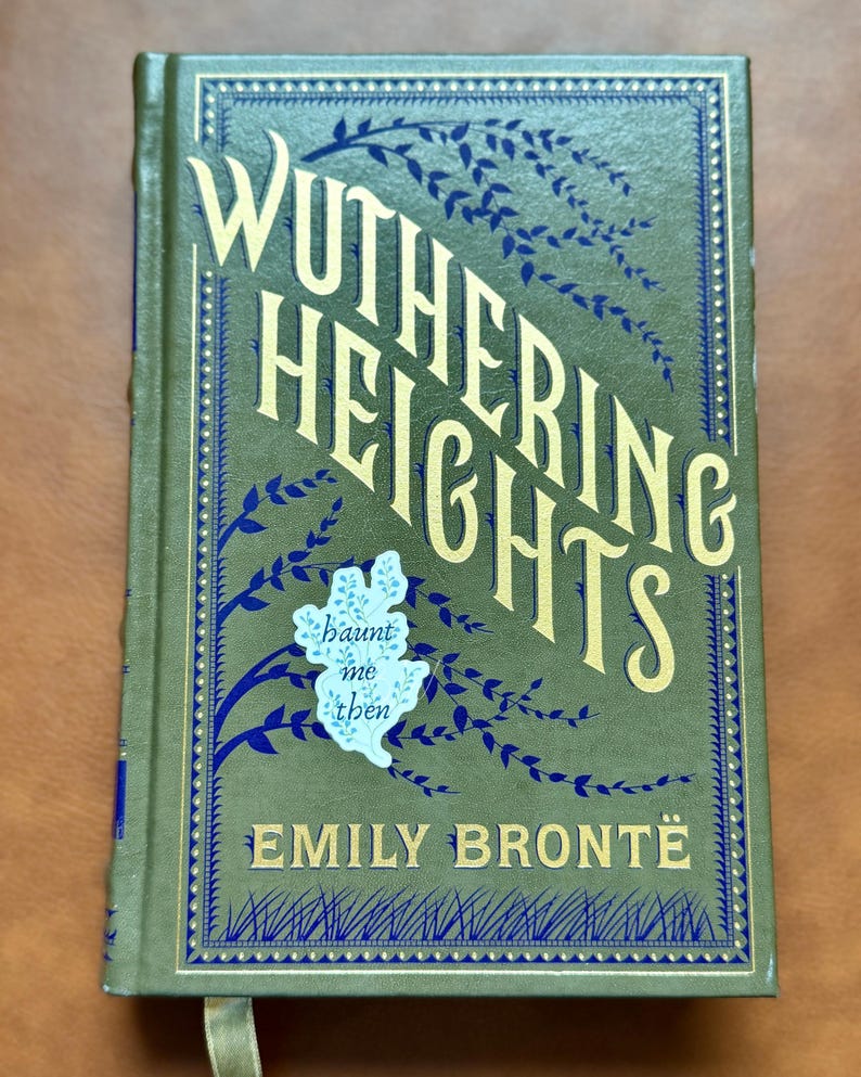 K&ouml;nnte beinhalten: Ein Hardcover-Buch mit dem Titel "Wuthering Heights" von Emily Bront&euml;. Das Cover ist olivgr&uuml;n mit goldfarbenen Buchstaben und blauen floralen Akzenten. Ein kleiner Aufkleber mit den Worten "haunt me then" befindet sich auf dem Cover.
