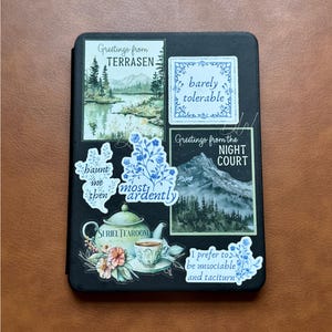 K&ouml;nnte beinhalten: Ein schwarzes Notizbuch, das mit verschiedenen Aufklebern verziert ist. Die Aufkleber zeigen Landschaftsbilder, florale Muster und S&auml;tze wie "Greetings from Terrasen", "barely tolerable" und "Suriel Tearoom". Die Aufkleber sind in Blau, Gr&uuml;n und Wei&szlig; gehalten.