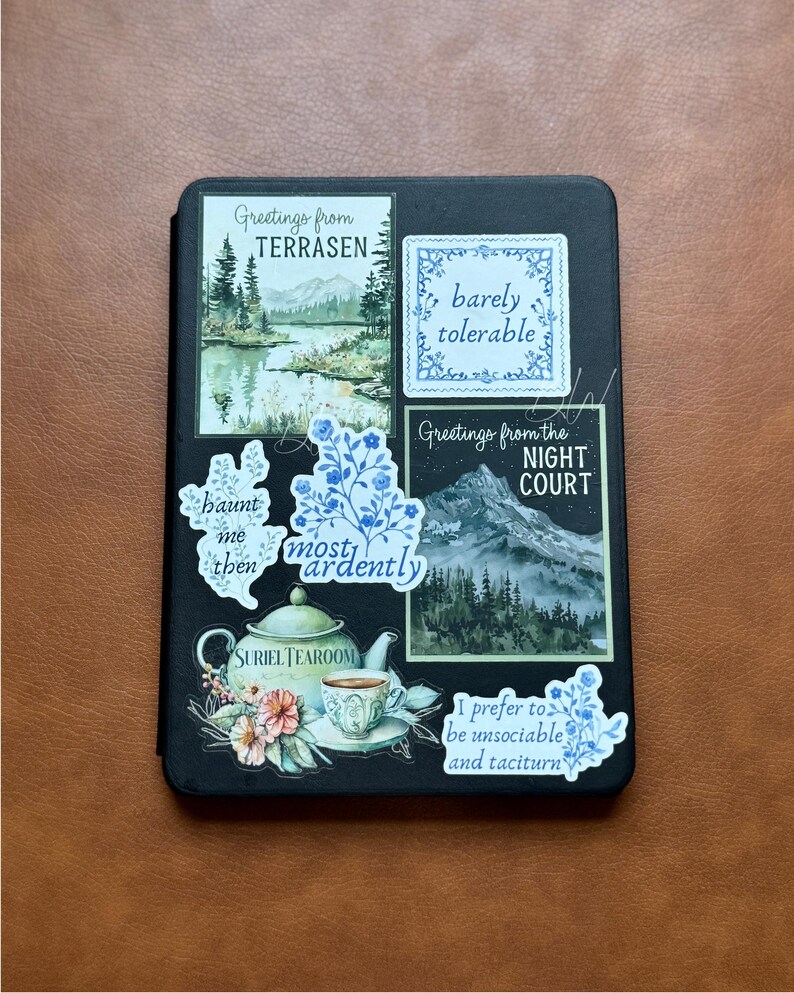 K&ouml;nnte beinhalten: Ein schwarzes Notizbuch, das mit verschiedenen Aufklebern verziert ist. Die Aufkleber zeigen Landschaftsbilder, florale Muster und S&auml;tze wie "Greetings from Terrasen", "barely tolerable" und "Suriel Tearoom". Die Aufkleber sind in Blau, Gr&uuml;n und Wei&szlig; gehalten.